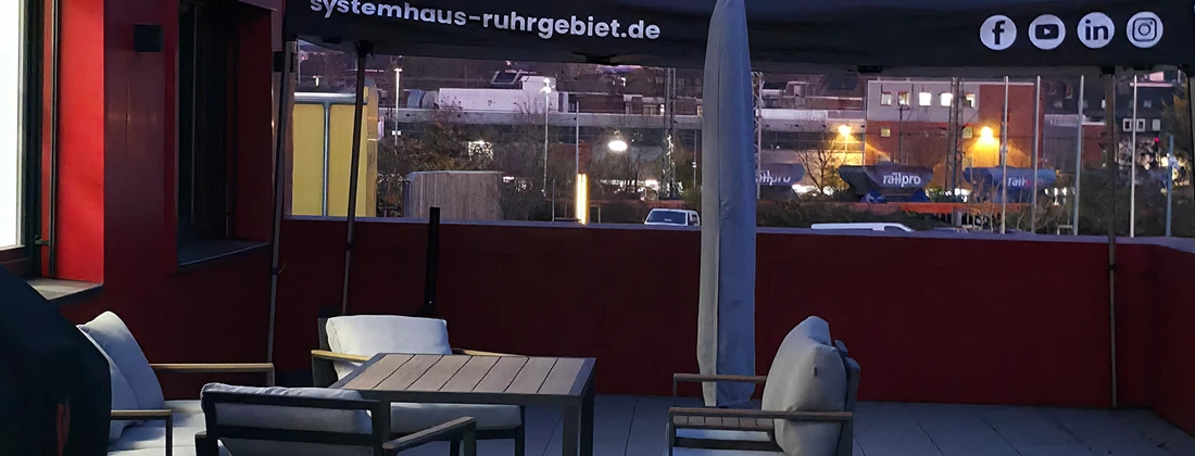 Terrasse mit Sitzmöbeln und einem Sonnenschirm, der das Logo von systemhaus-ruhrgebiet.de zeigt.
