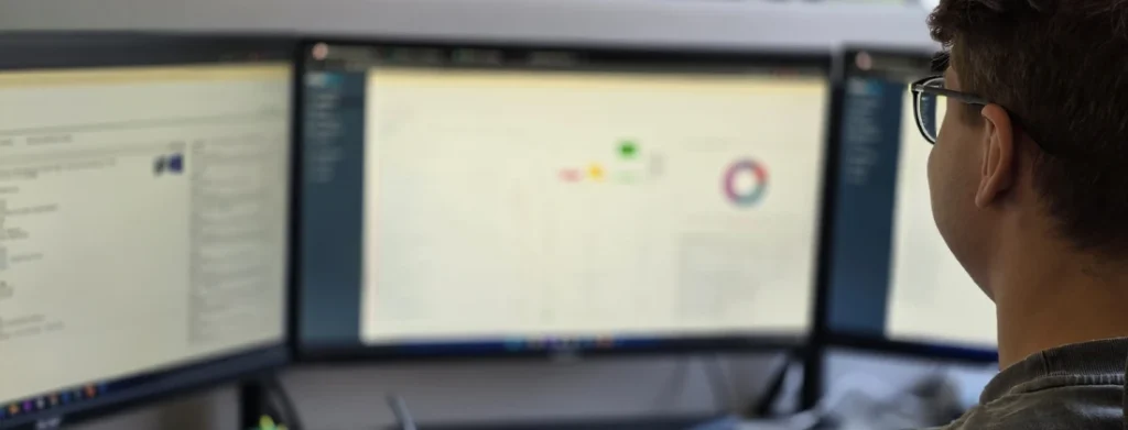 Person mit Brille sitzt vor drei Monitoren, die Datenvisualisierungen und Softwareanwendungen anzeigen.