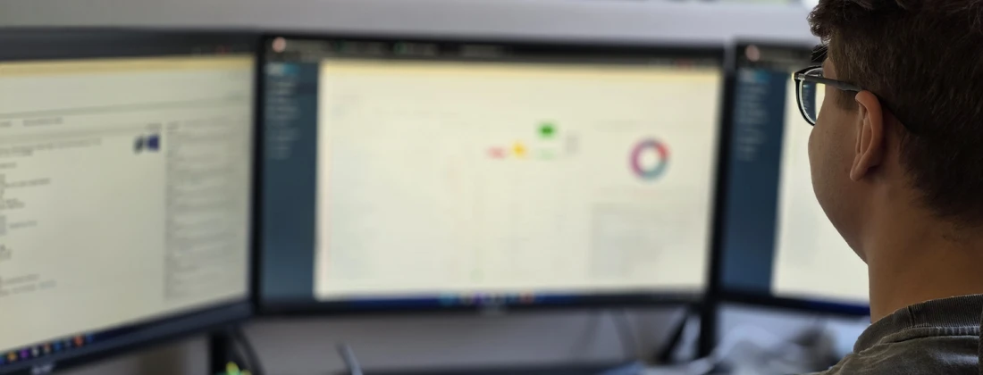Person mit Brille sitzt vor drei Monitoren, die Datenvisualisierungen und Softwareanwendungen anzeigen.
