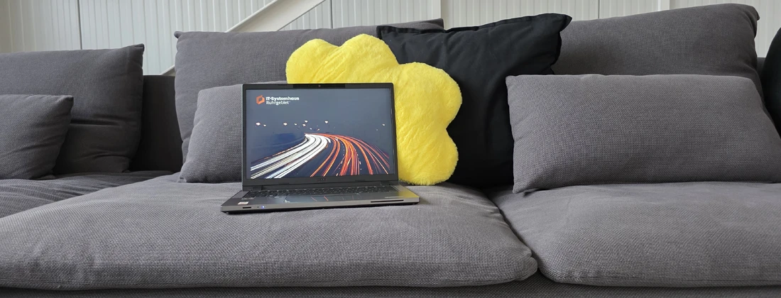 Laptop auf einem grauen Sofa mit einem gelben Kissen und einer schwarzen Kissen im Hintergrund.