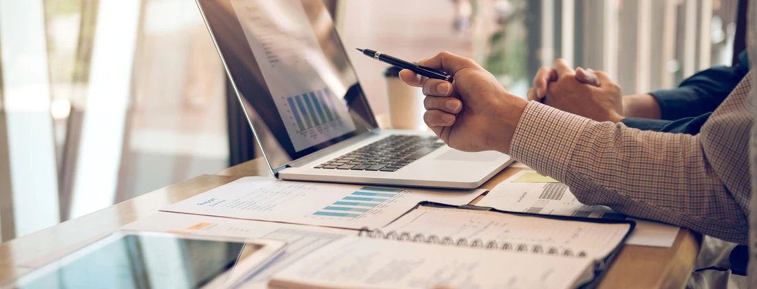 Hände, die auf einen Laptop mit Diagrammen und Statistiken zeigen, während Notizen und ein Tablet auf dem Tisch liegen.