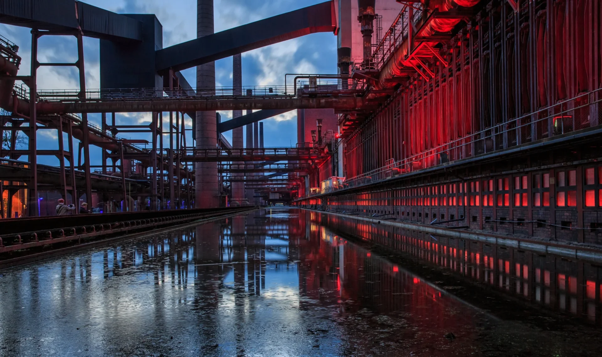 Industrielle Anlage mit reflektierendem Wasser und roten Lichtern in der Dämmerung.