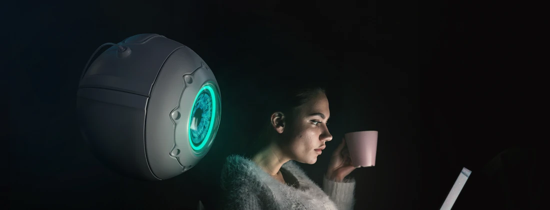 Eine Frau sitzt mit einer Tasse in der Hand vor einem Laptop, während ein futuristisches Gerät im Hintergrund leuchtet.