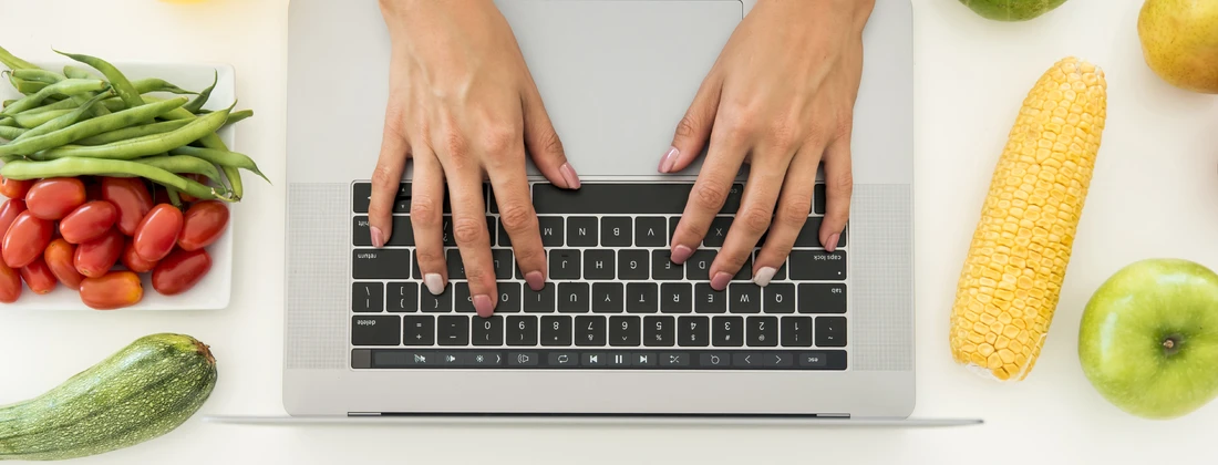 Hände tippen auf einer Laptop-Tastatur, umgeben von frischem Gemüse und Obst wie Tomaten, Bohnen, Mais und Äpfeln.