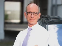 Frank Schumacher in einem weißen Hemd und einer hellen Krawatte, lächelnd vor einem Fahrzeug.