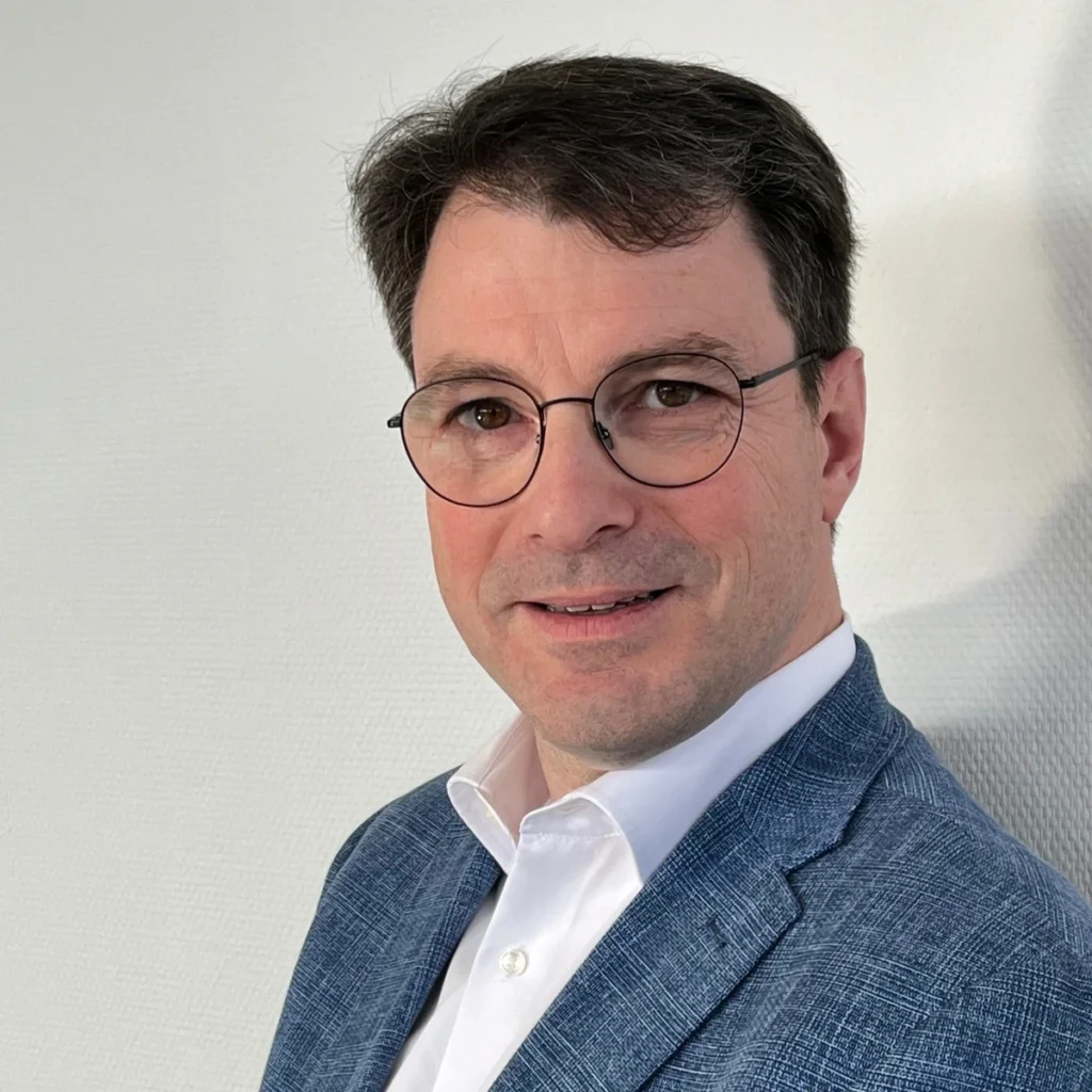 Mann mit Brille und Anzug, der in die Kamera lächelt, vor einer hellen Wand.