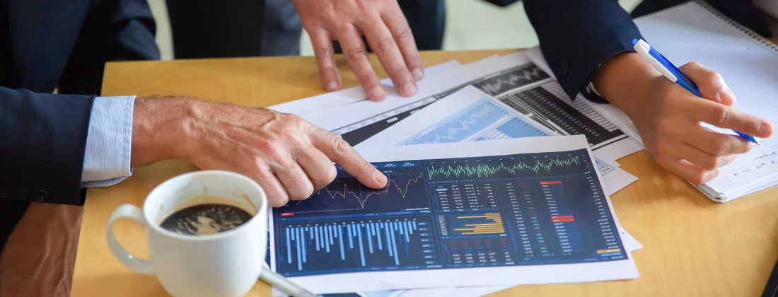 Hände, die auf Finanzdiagramme und Datenanalysen auf einem Tisch zeigen, mit einer Tasse Kaffee im Vordergrund.