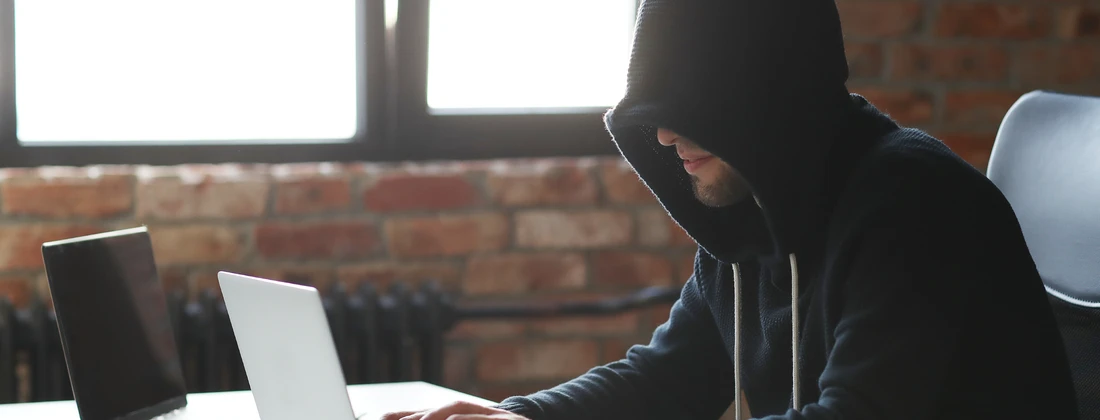 Person mit Kapuze, die an einem Laptop in einem Raum mit Backsteinwand arbeitet.