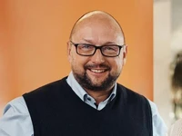 Manuel Hübner mit Brille und Weste lächelt in die Kamera vor einem orangefarbenen Hintergrund.