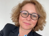 Nicole Ruschke mit lockigem Haar und Brille, in einem schwarzen Blazer, lächelt in die Kamera.