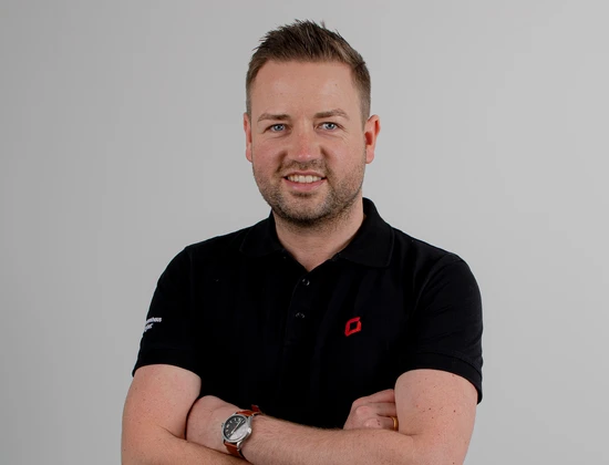 Nils Kathagen mit verschränkten Armen in einem schwarzen Poloshirt mit Logo auf grauem Hintergrund.