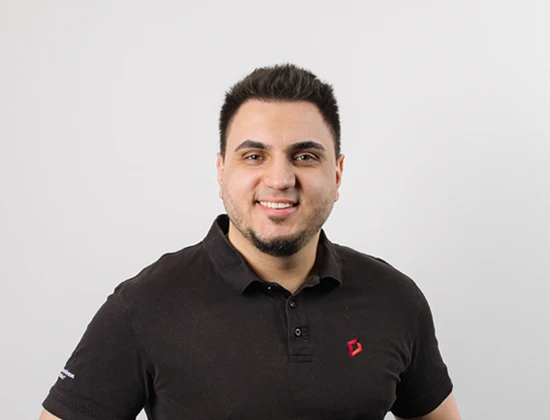 Emir T in einem schwarzen Polohemd mit einem lächelnden Gesichtsausdruck vor einem neutralen Hintergrund.