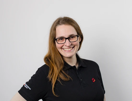 Jacqueline P. mit langen Haaren und Brille, trägt ein schwarzes Polohemd mit Logo.