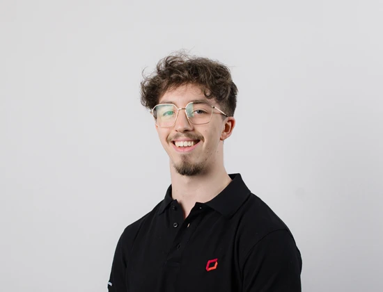 Nick A. mit Brille und lockigem Haar in schwarzem Poloshirt, lächelnd vor neutralem Hintergrund.
