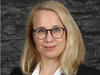 Simone Jegus, eine Fachkraft im Bereich Medizintechnik, mit blonden Haaren und Brille vor einer dunklen Steinwand.