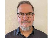 Uwe Ulrich mit Brille und grauem Bart, in einem hellen Hemd vor einer neutralen Wand.