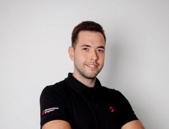 Florian in einem schwarzen Poloshirt mit Logo, lächelt und steht mit verschränkten Armen vor einem neutralen Hintergrund.