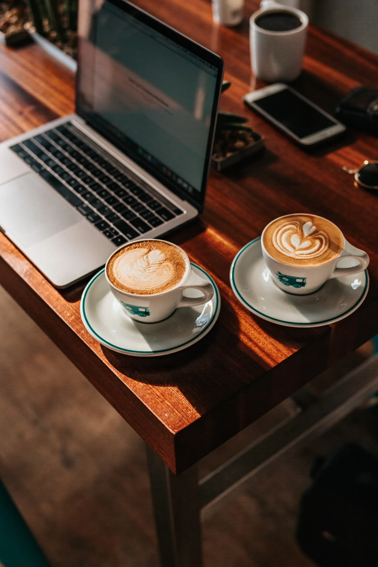 Zwei Tassen Kaffee mit Latte-Art auf einem Holztisch neben einem Laptop und einem Smartphone.