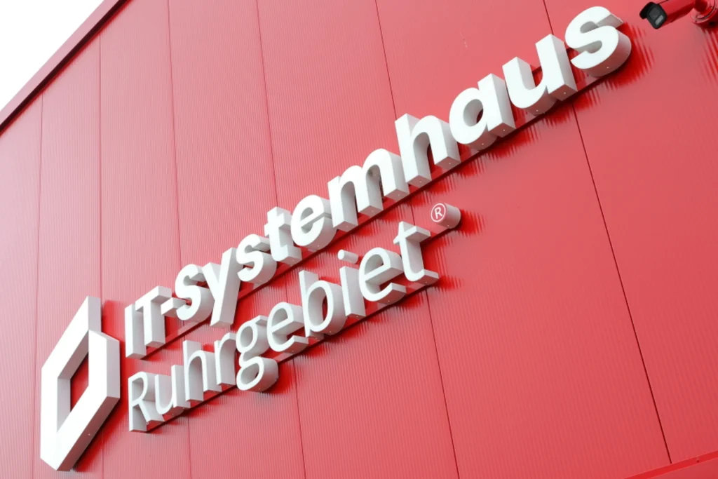 Rotes Schild mit der Aufschrift 'IT-Systemhaus Ruhrgebiet' in moderner Schriftart an einer Wand.